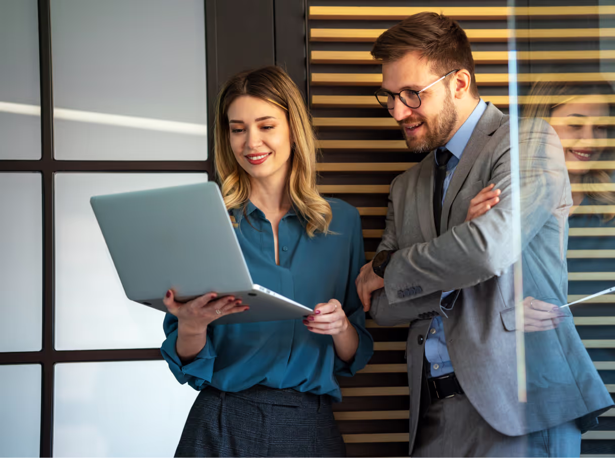 Two business professionals reviewing information on a laptop together in a modern office space with wood-slatted wall features. One wearing a teal blouse and the other in a grey suit jacket with glasses, both smiling while looking at the screen